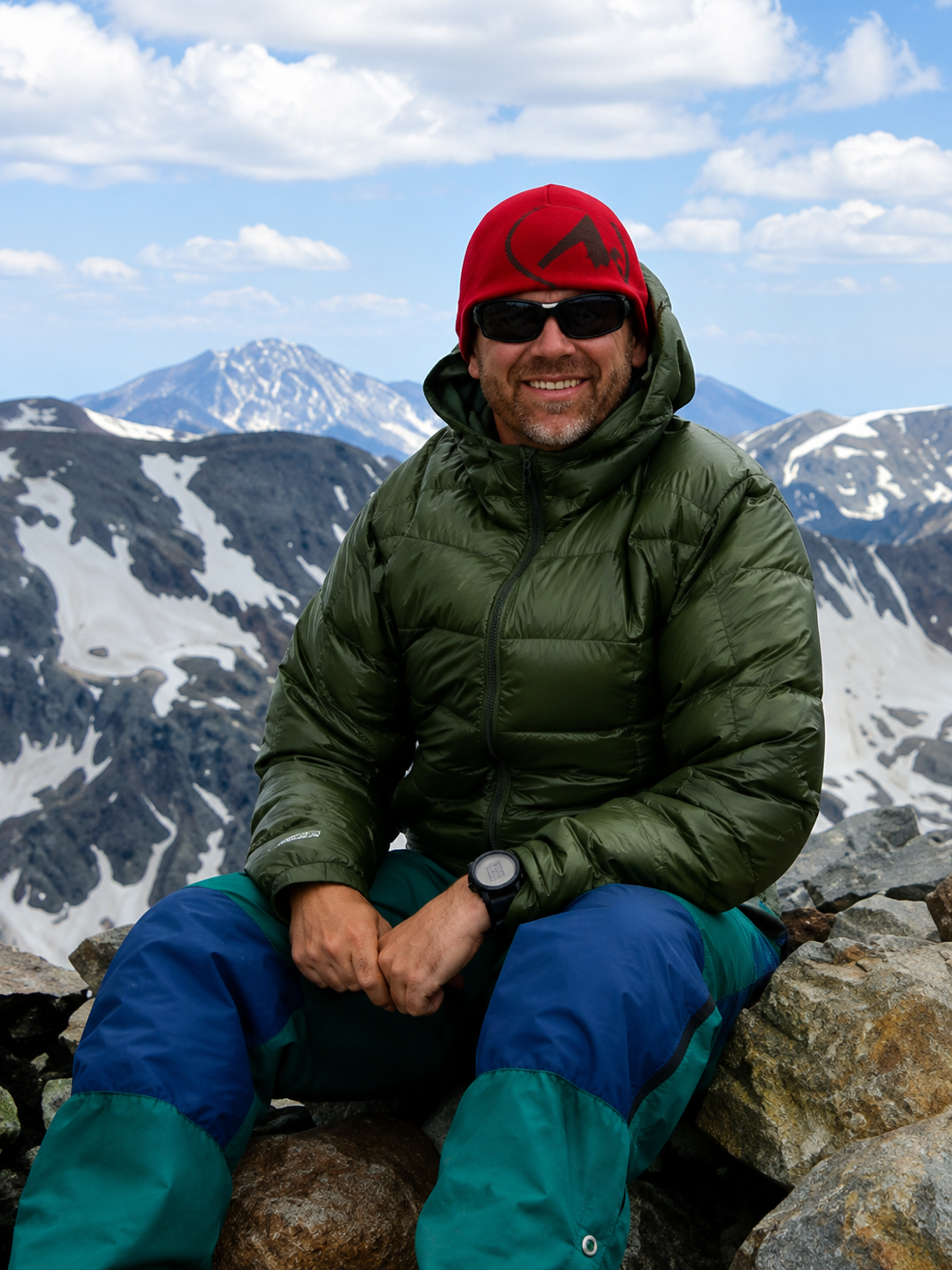 Smiling climber in red helmet and green jacket sitting on rocks with snowy mountains behind