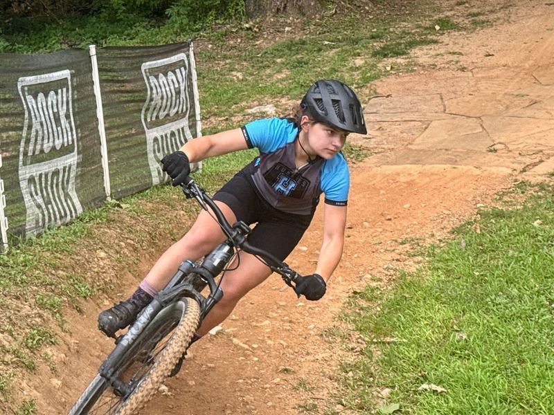Mountain biker in blue shirt leans into a dirt turn on a trail, wearing a helmet and gloves.