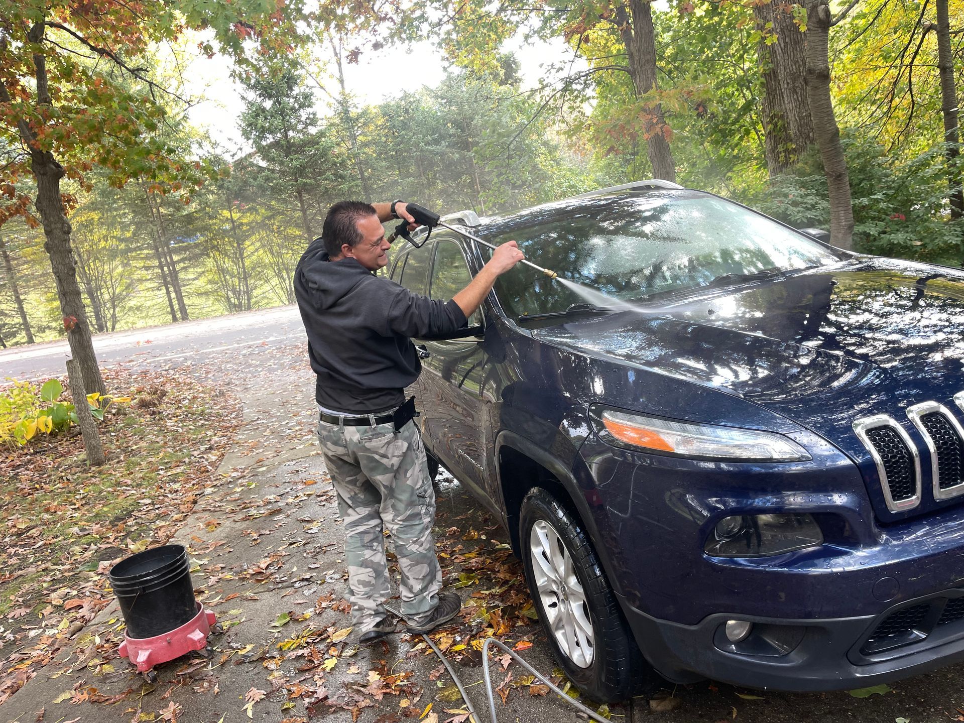 Man washing a dark blue SUV with a hose on a leafy roadside in autumn