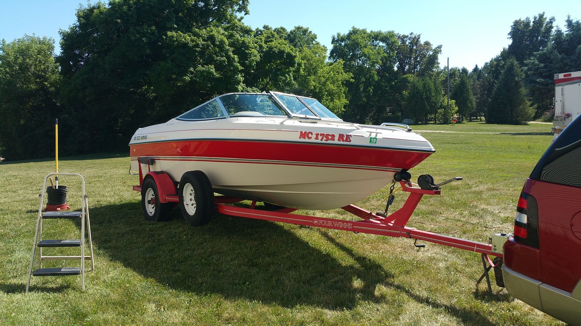 White and red boat on trailer hitched to red SUV in a grassy field, with a step ladder nearby