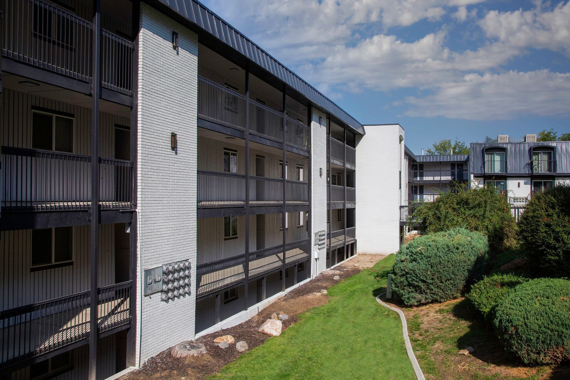 Apartment complex exterior with balconies, grass, and blue sky with clouds.