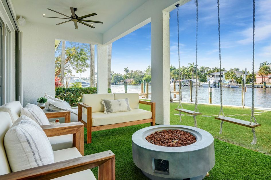 Covered patio with white chairs, fire pit, hanging swing, and waterfront view through open columns