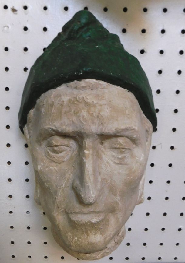 White carved face sculpture wearing a dark green pointed cap, displayed on a pegboard background