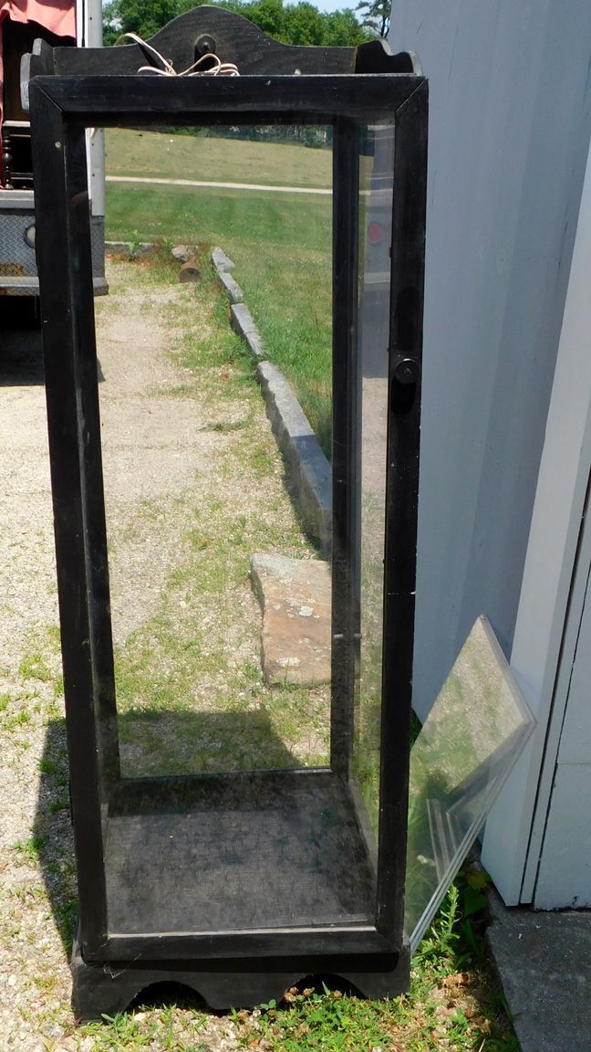Black-framed glass display cabinet standing outdoors on grass and concrete beside a white building.