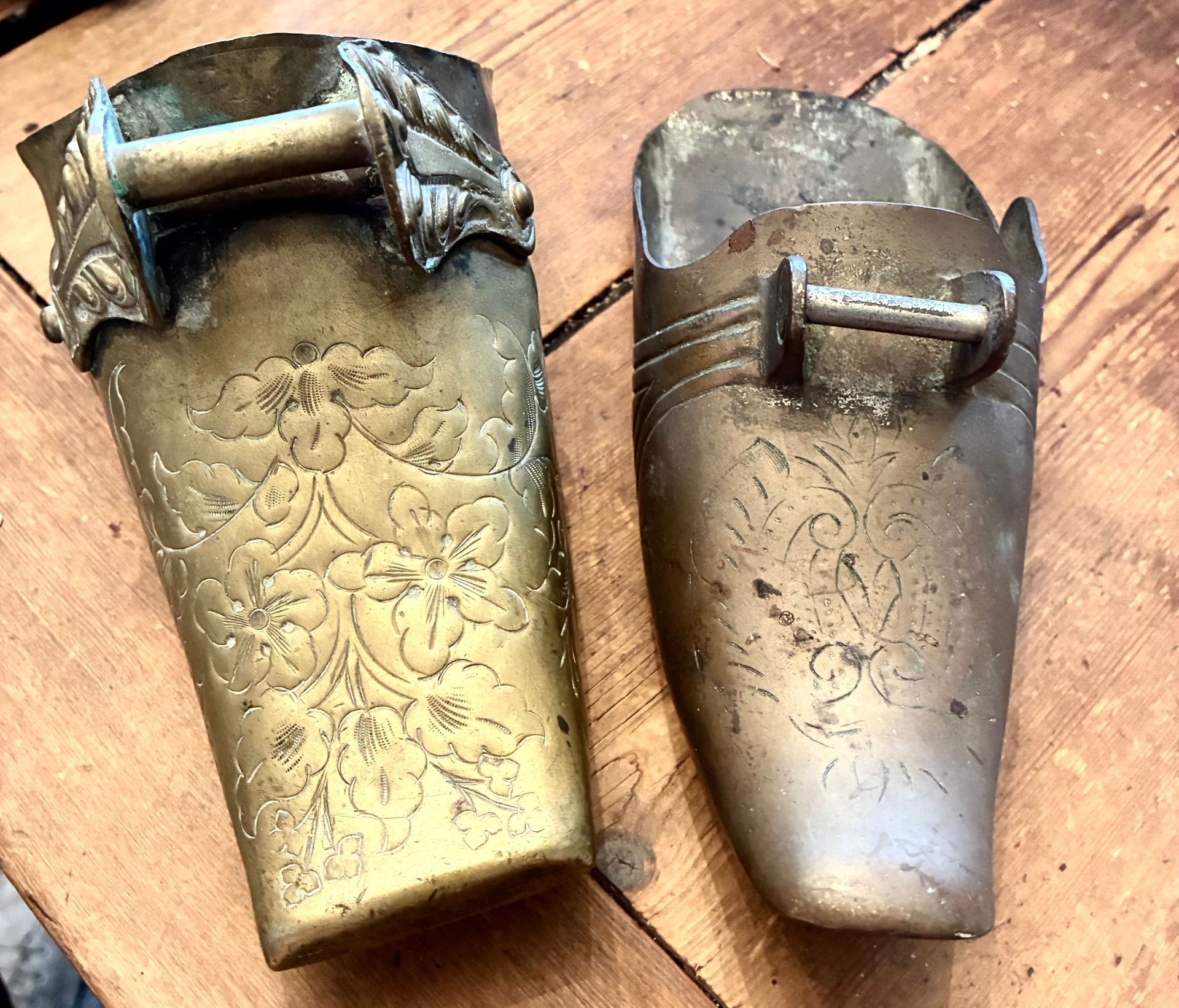 Two ornate brass cuffs with floral engraving and metal handles on a wooden surface