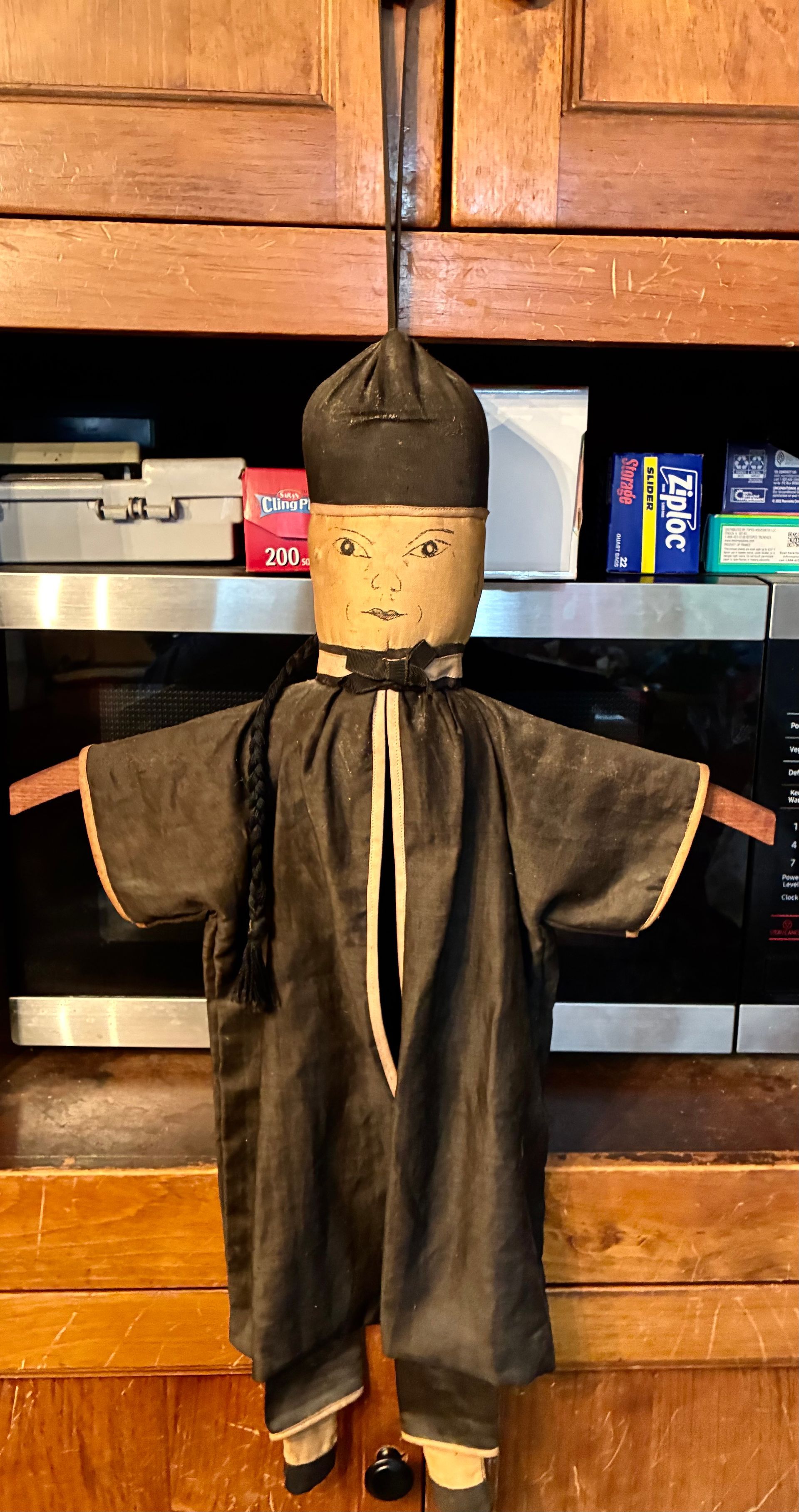 Small handmade doll in dark robes with a pointed hat, standing on a shelf in front of wood cabinets.