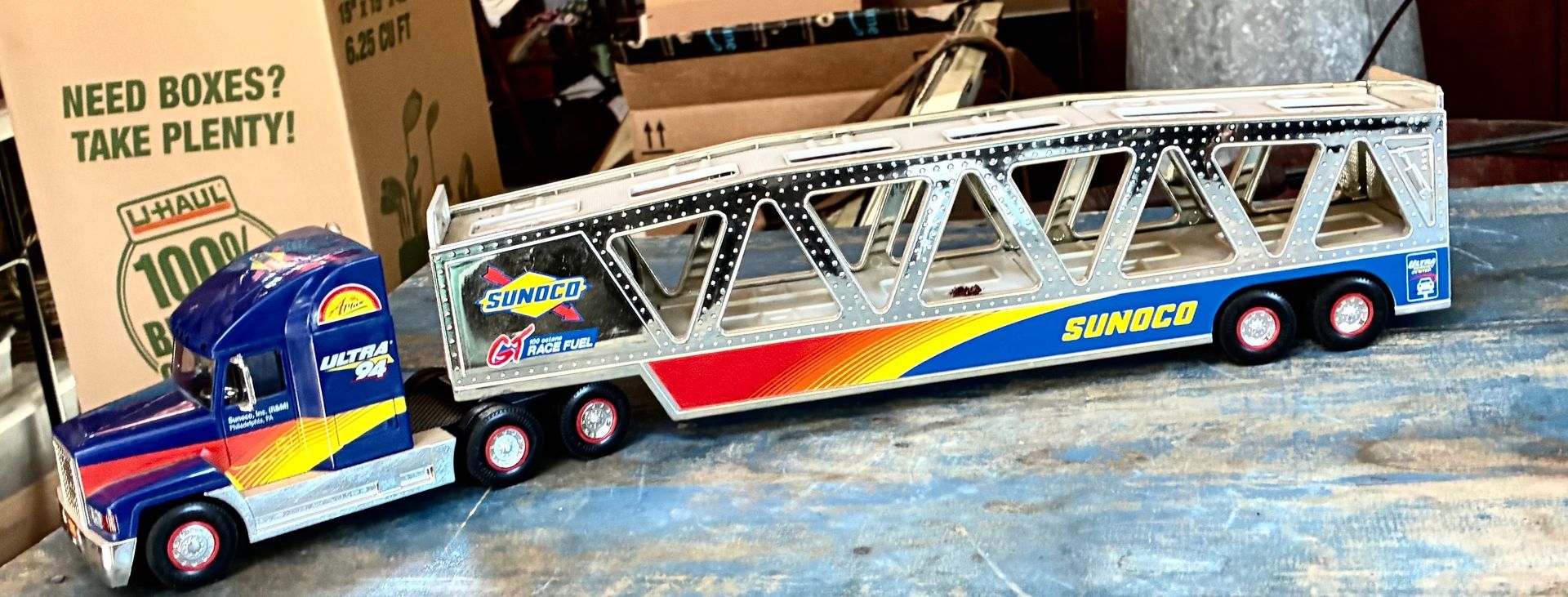 Toy race car hauler trailer with colorful graphics parked beside a cardboard sign reading “Need boxes? Take plenty!”
