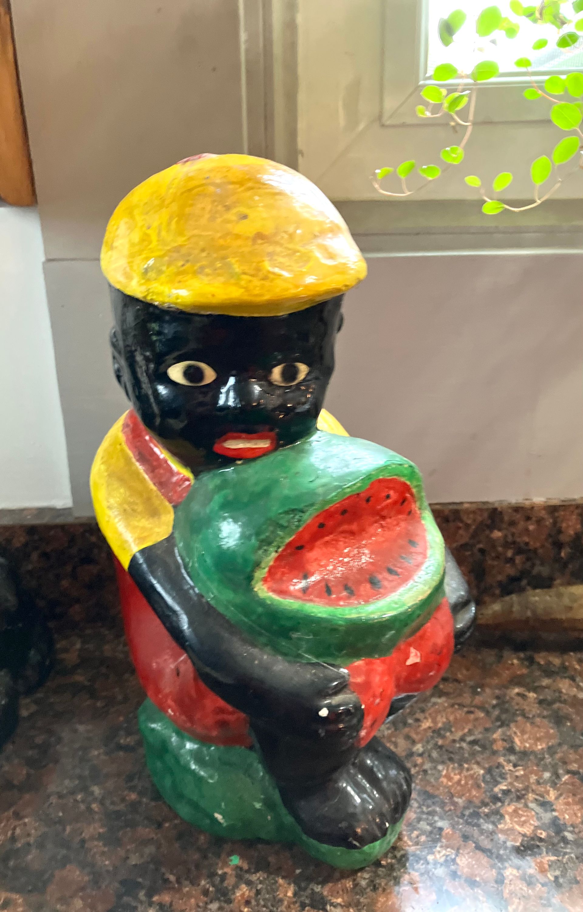 Colorful painted figurine of a child holding a green-and-red bowl, wearing a yellow hat indoors