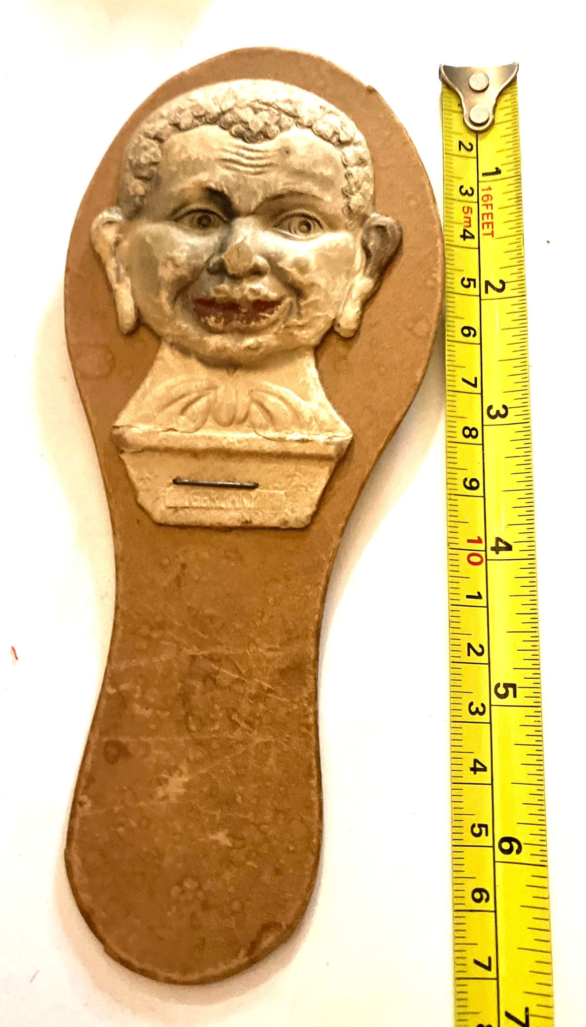 Brown sole-shaped plaque with a small sculpted face at the top, shown beside a yellow measuring tape.