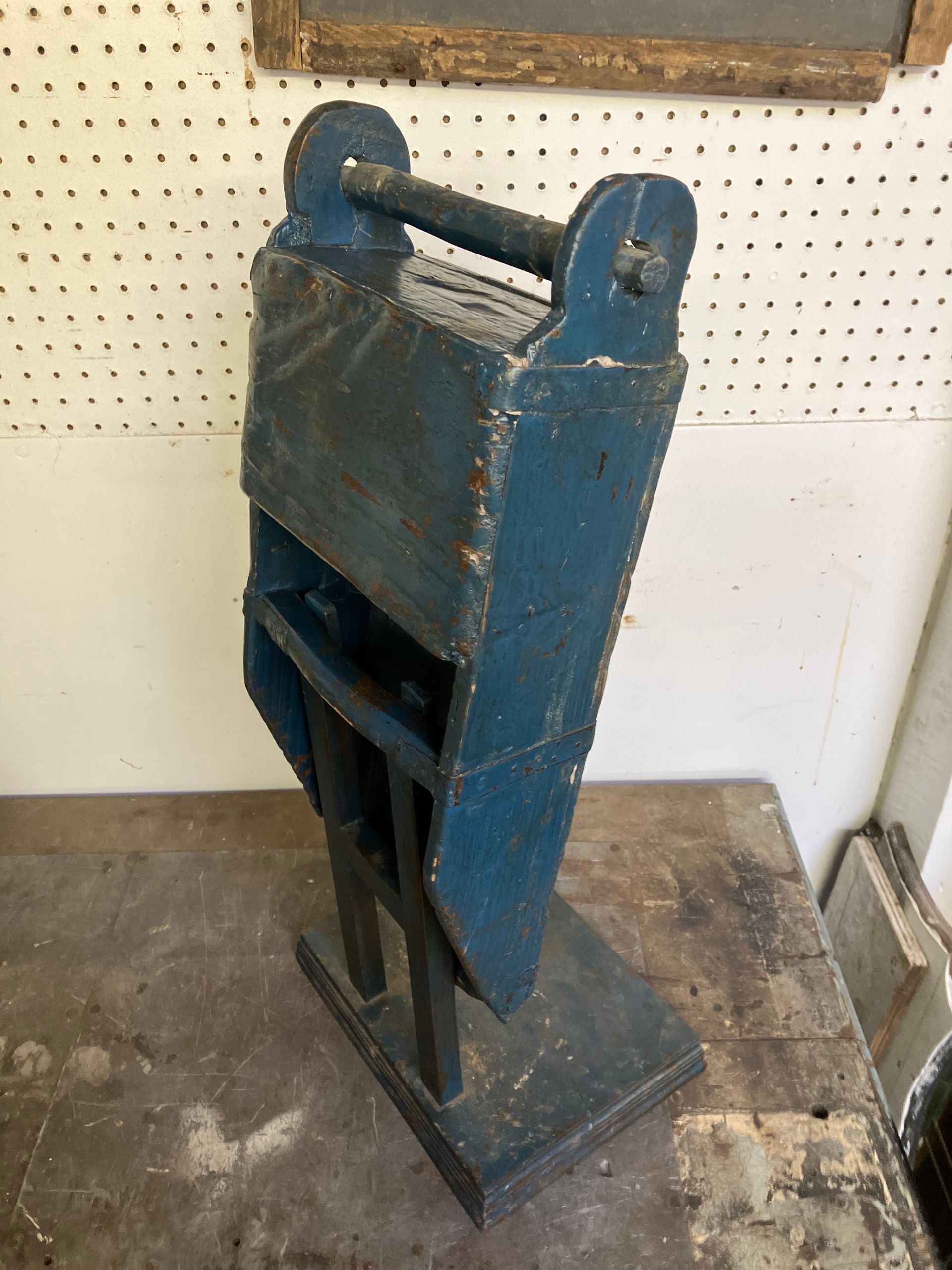 Blue metal paper cutter on a stand in a workshop, against a pegboard wall.