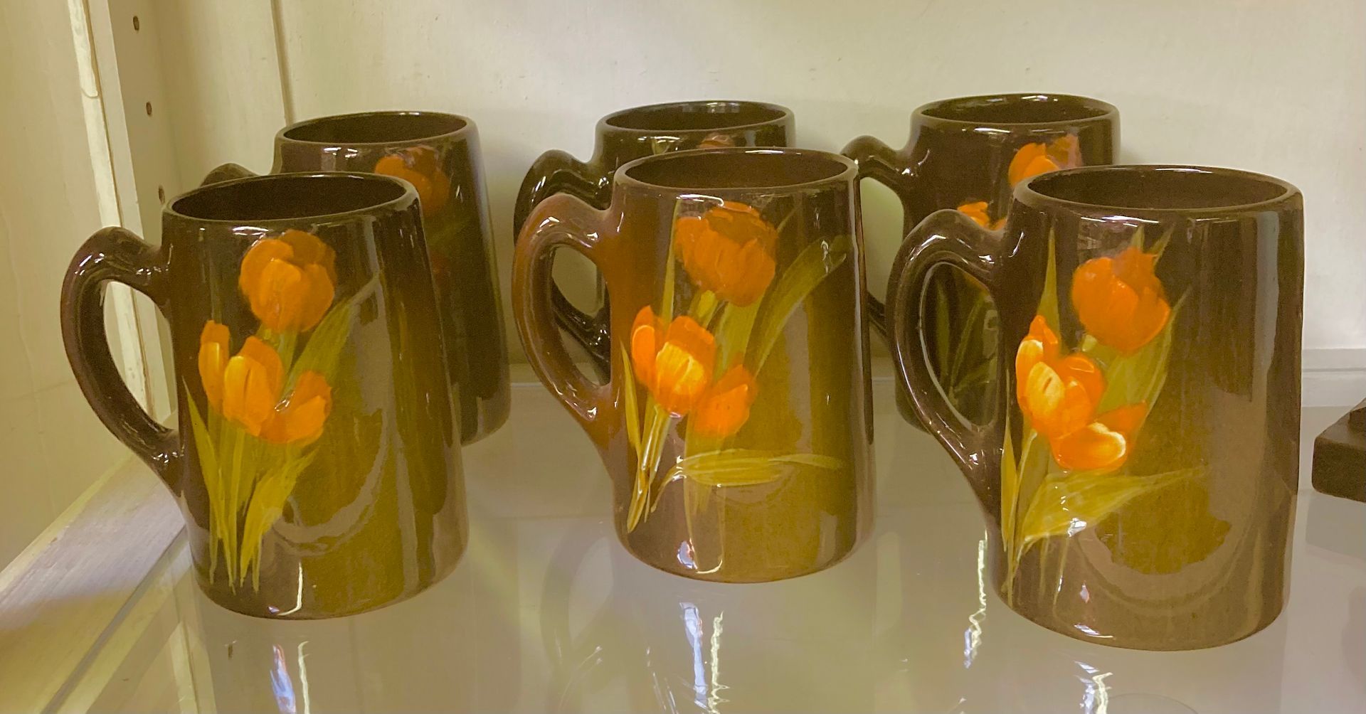 Five dark glass mugs with orange fruit and flower designs, lined up on a reflective surface.