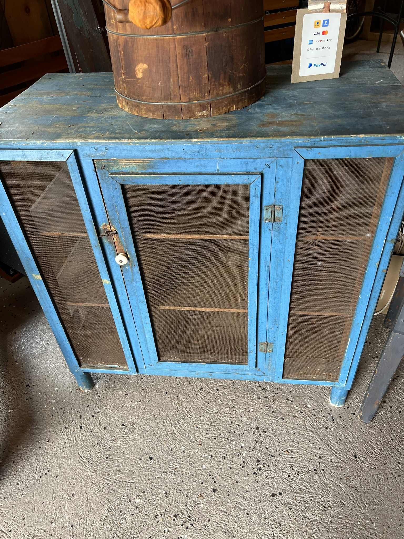 Blue wooden cabinet with wire-mesh doors beneath a barrel, on a gravel floor.