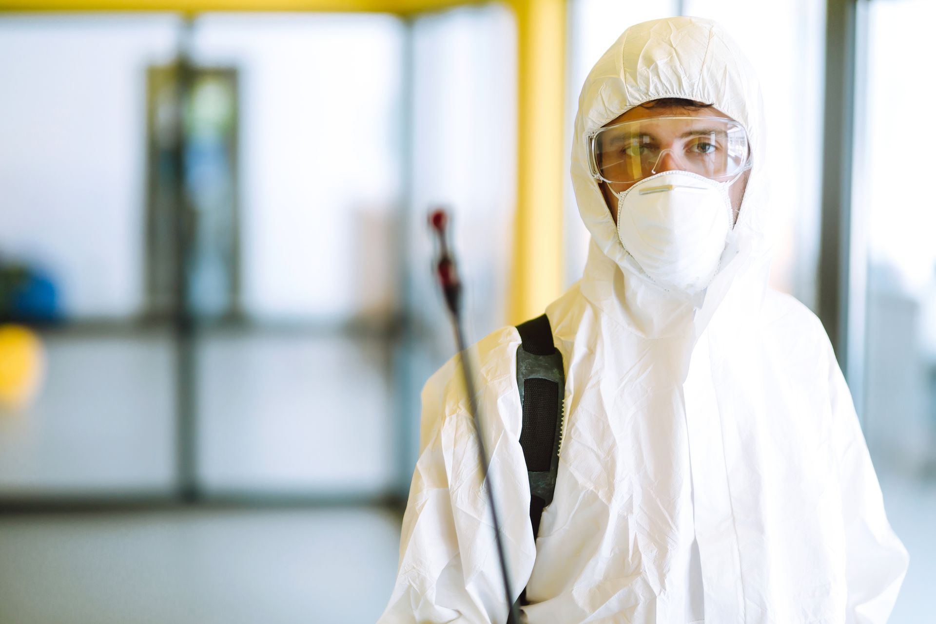 Person in white protective suit, mask, and goggles standing in a bright hallway.