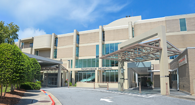 Exterior of a beige hospital building with glass entrance and covered drop-off canopy under a blue sky