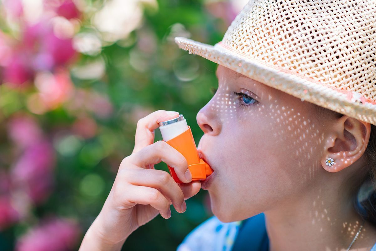 Child using an orange inhaler outdoors, wearing a straw hat, with pink flowers in the background