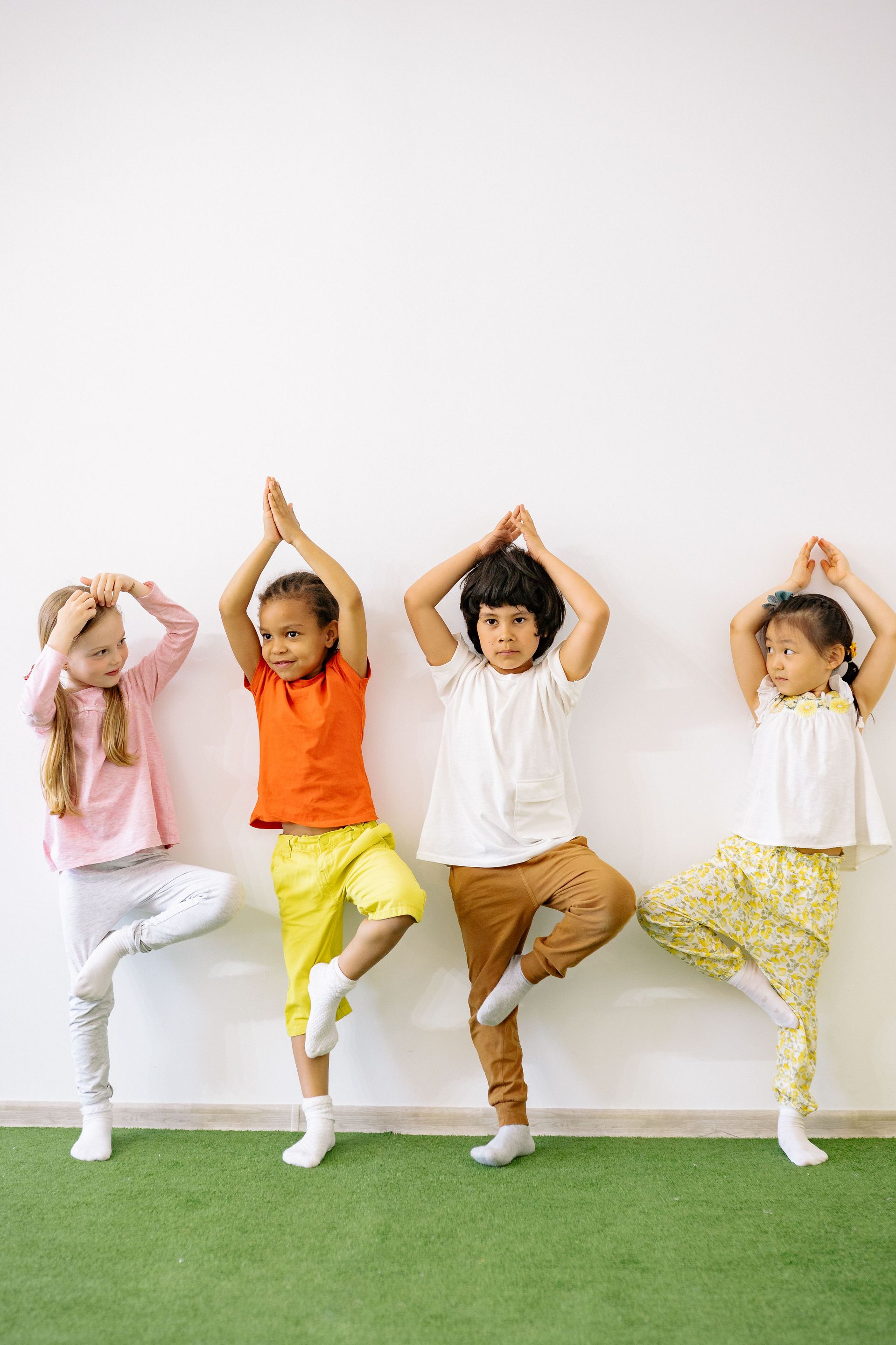 Four children in colorful clothes pose with arms raised against a white wall and green floor.