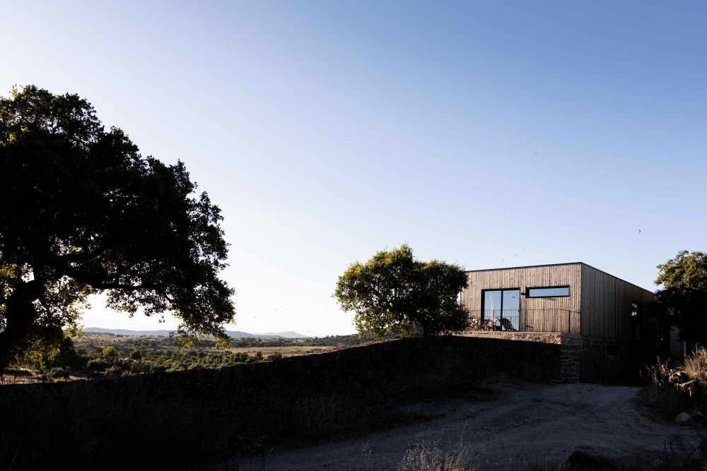 Wooden cabin nestled in a rural landscape, framed by trees and a stone wall. Clear sky overhead.