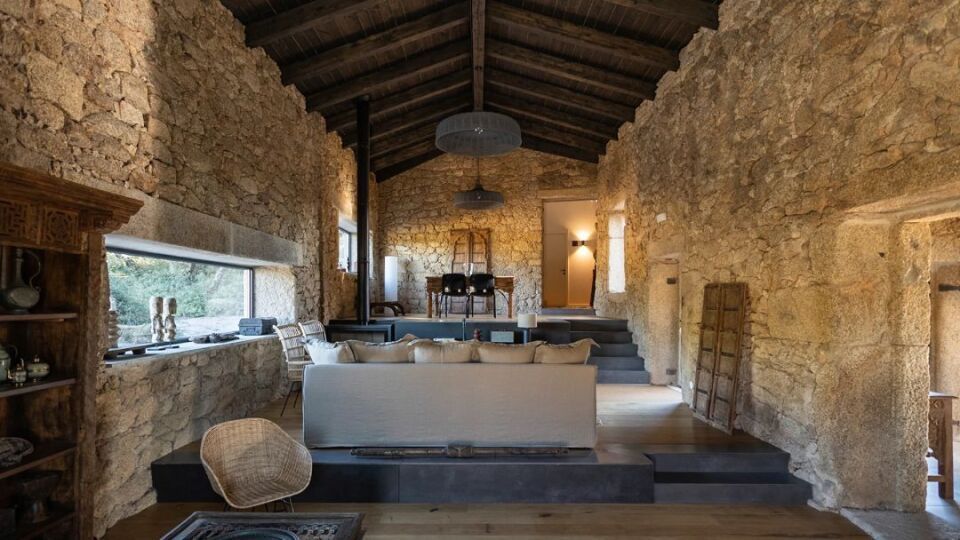 Stone-walled interior with a couch, fireplace, and exposed wood ceiling. Natural light streams from the windows.