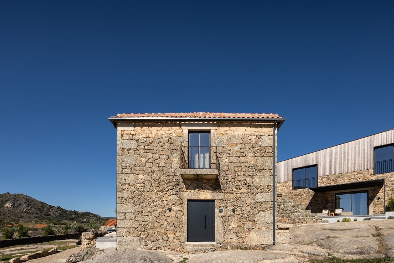 Stone building with dark door and balcony under a blue sky, connected to a modern building with wood panels.