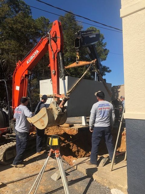 Workers with an excavator lifting a concrete slab near a building, with a tripod in the foreground.