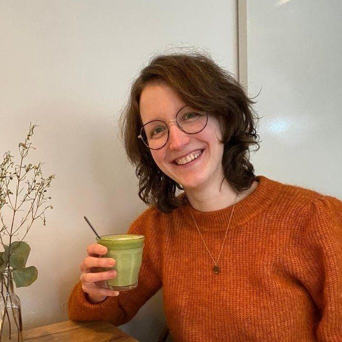 Een lachend persoon in een oranje trui houdt een groene beker vast naast een vaas met gedroogde bloemen.