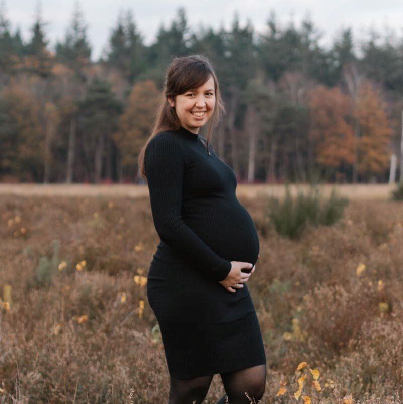 Een zwangere vrouw in een zwarte jurk staat in een grasveld en lacht naar de camera.