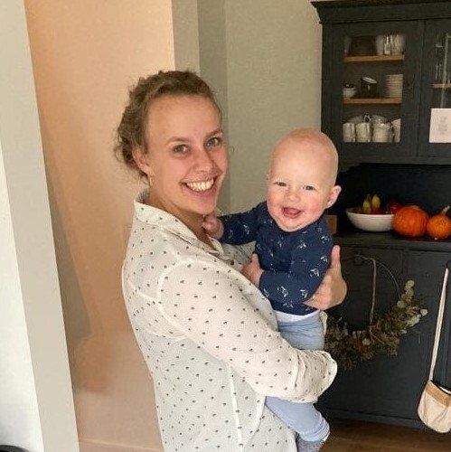 Een lachende vrouw houdt een lachende baby vast in een keuken naast donkere keukenkastjes en een fruitschaal.