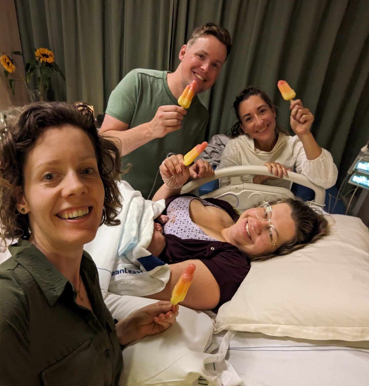 Vier lachende mensen in een ziekenkamer houden sinaasappelijsjes rond een patiënt die in bed ligt.