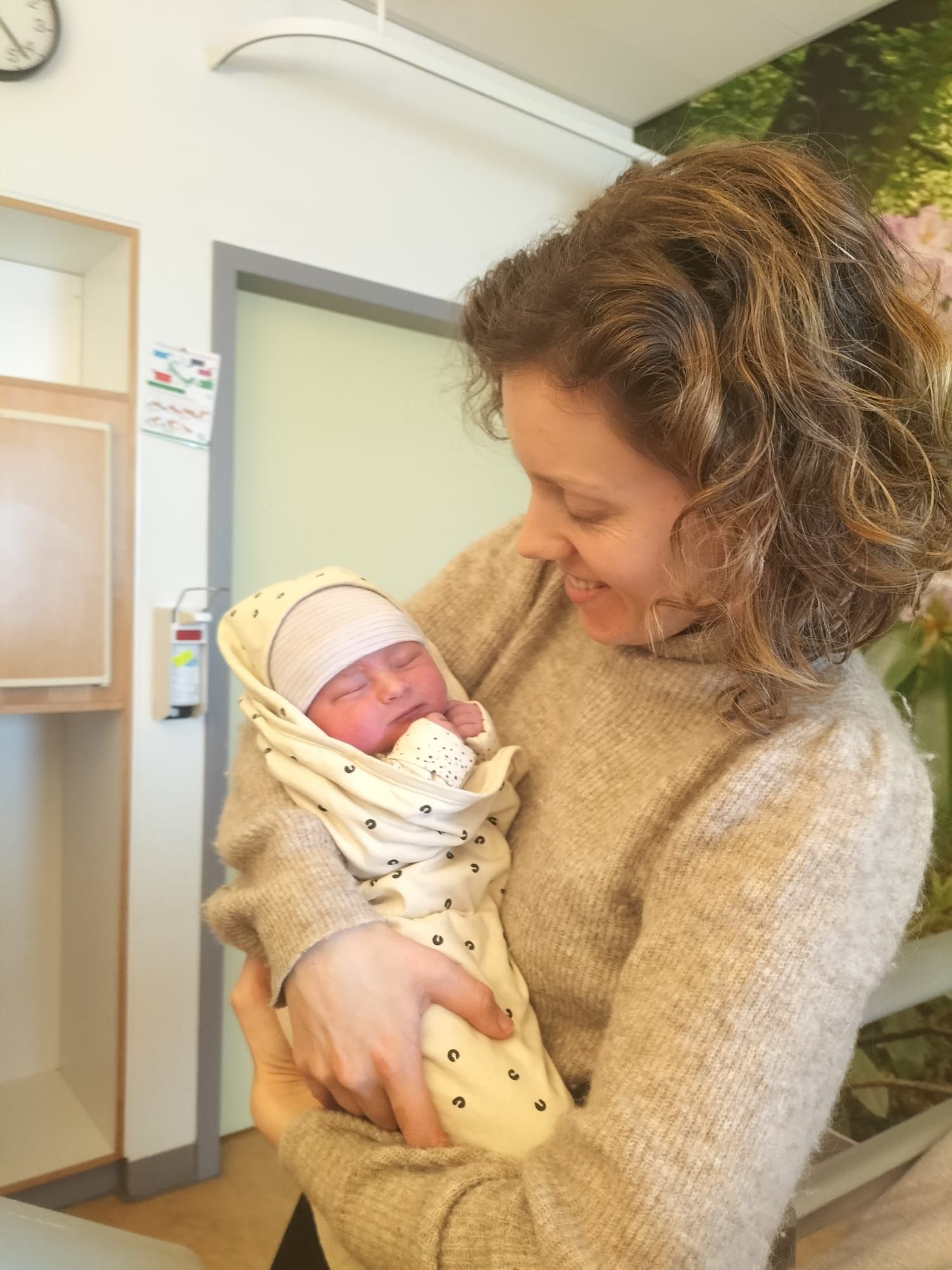 Een vrouw houdt een pasgeboren baby in een doek gewikkeld vast in een ziekenhuisgang en kijkt met een vriendelijke glimlach naar beneden.