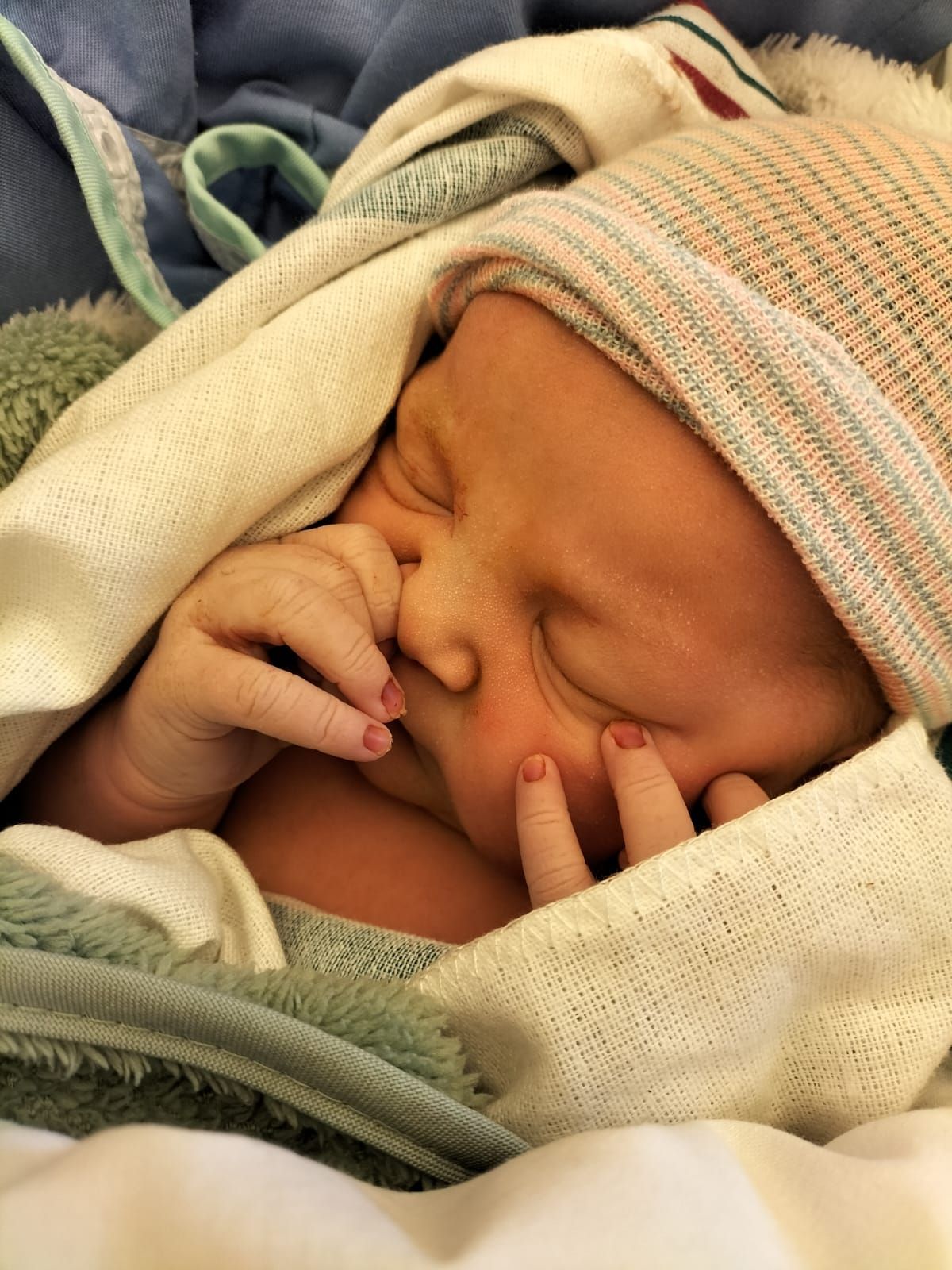 Een slapende baby, ingewikkeld in dekens, met de handjes dicht bij het gezicht en een gebreid mutsje over het hoofdje.