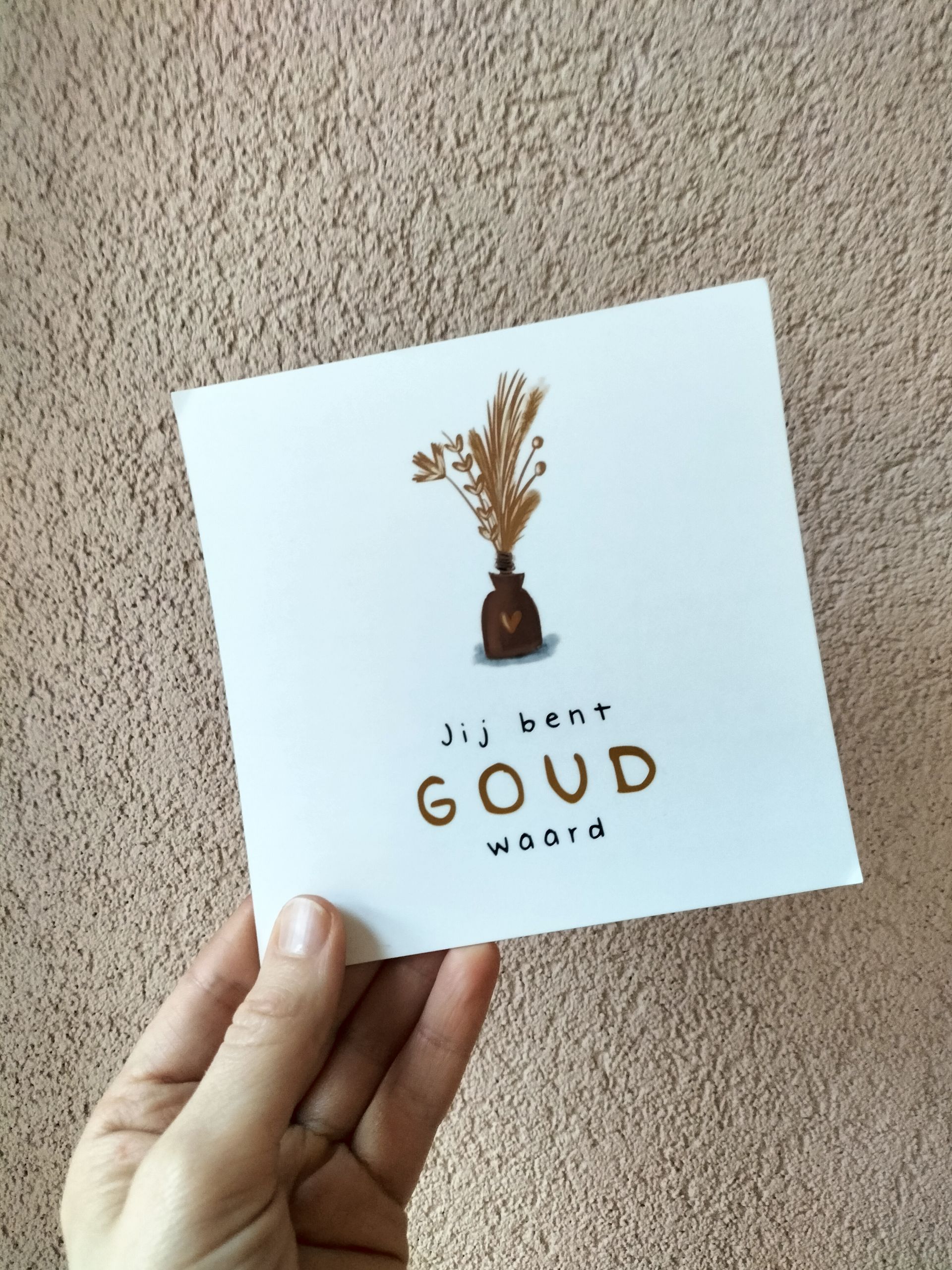 Een hand houdt een vierkante witte kaart vast met een gouden plantillustratie en de woorden