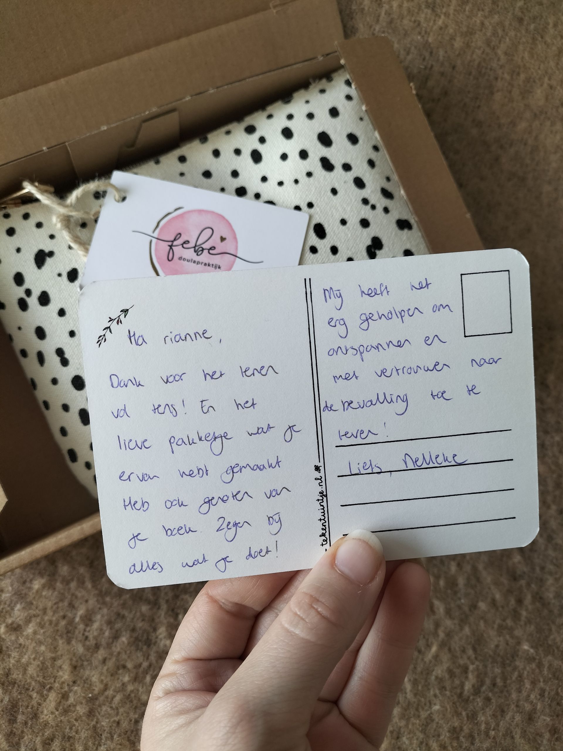 Een hand houdt een klein, handgeschreven bedankkaartje vast boven een geschenkdoos met stippen, gevuld met vloeipapier.