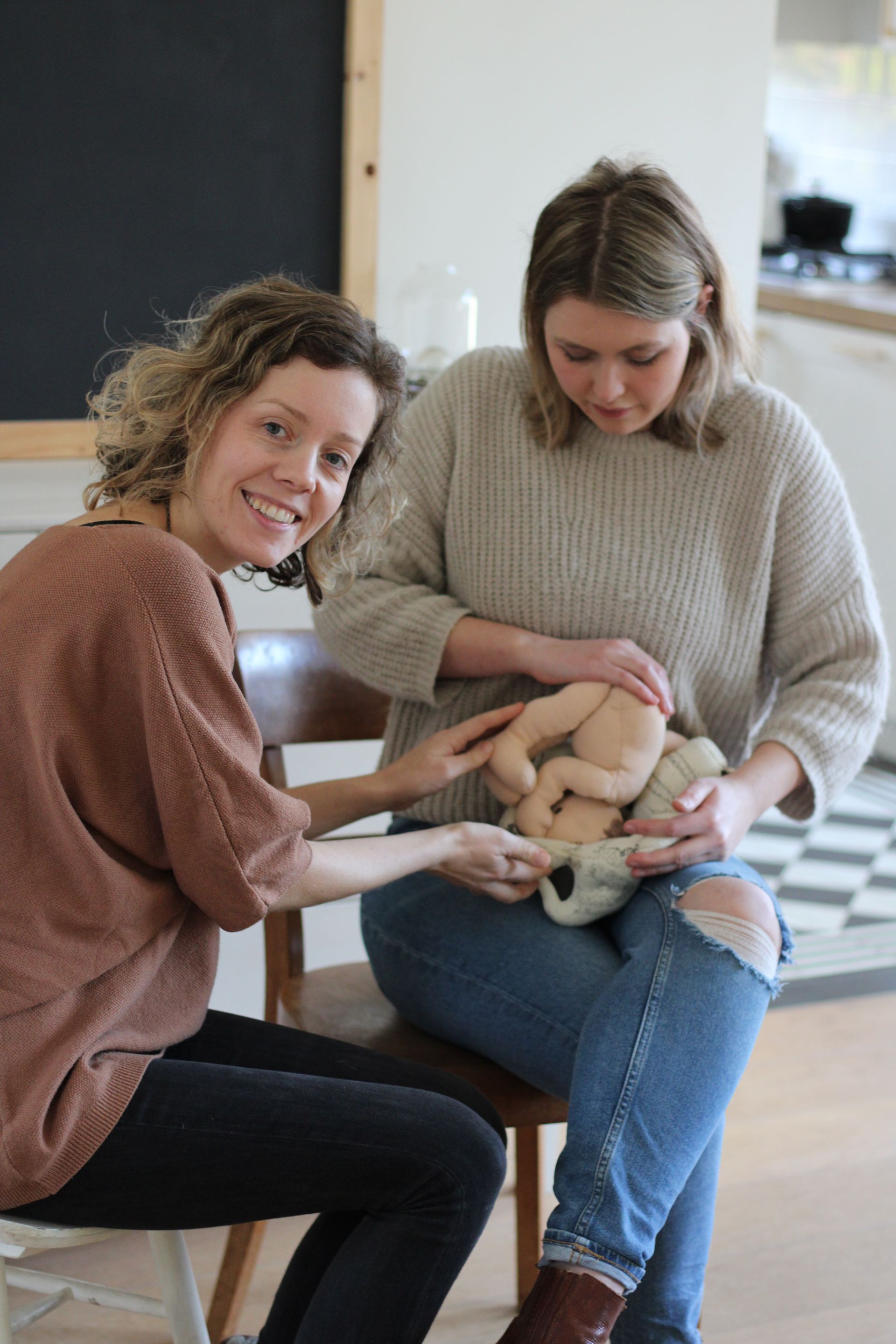 Twee vrouwen zitten op een bank en houden een pop met voorbeeldbekken vast, een van hen lacht naar de camera.
