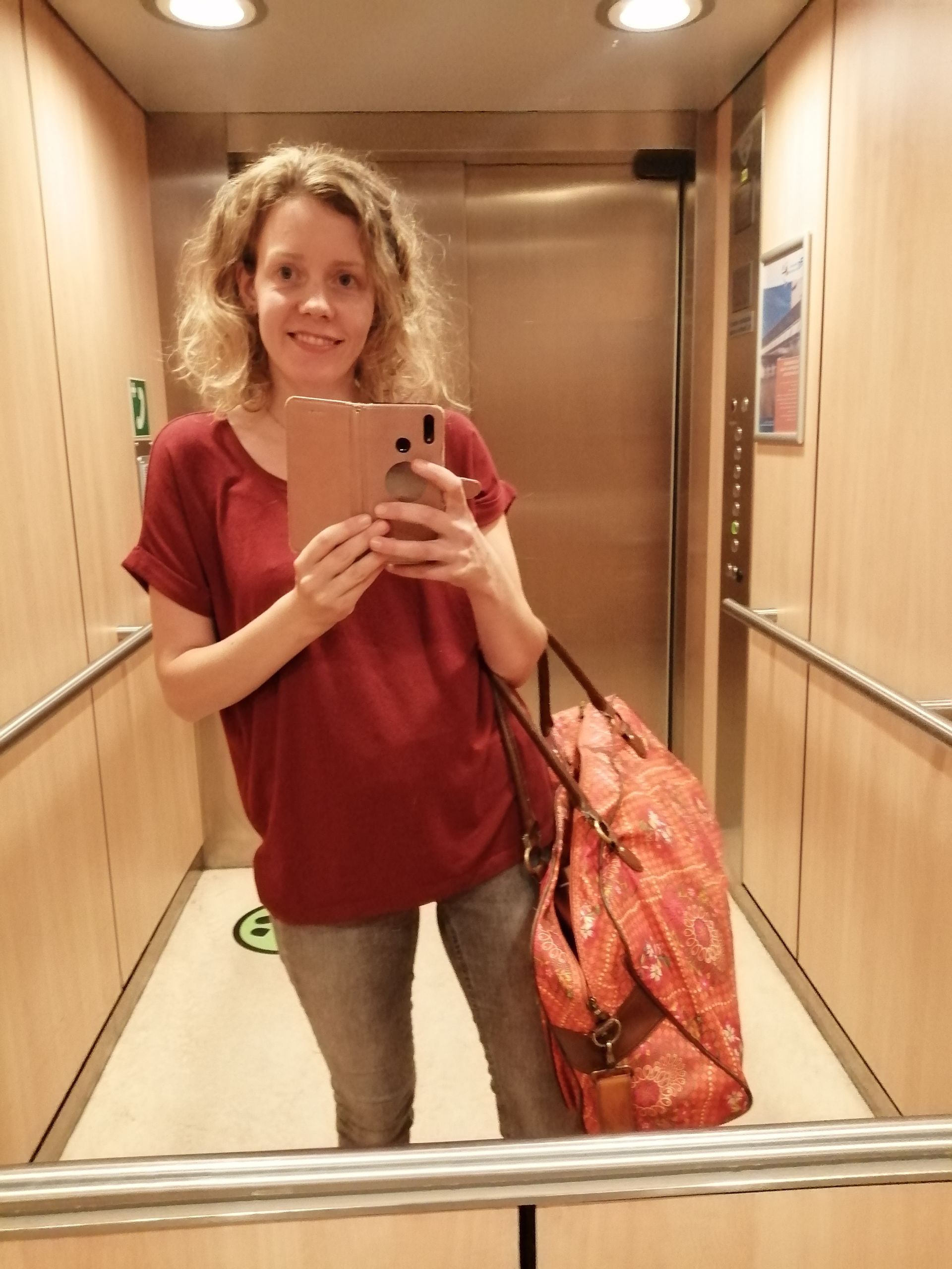 Een persoon die een selfie in de spiegel maakt in een lift, draagt een rood shirt en heeft een oranje tas bij zich.