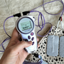 Een draagbaar paars elektronisch therapieapparaat in de hand van een persoon, met draden en elektroden op een bed.