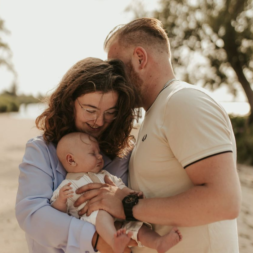 Een stel knuffelt een baby buiten in de warme zon, lachend en het kindje dicht tegen zich aan houdend.