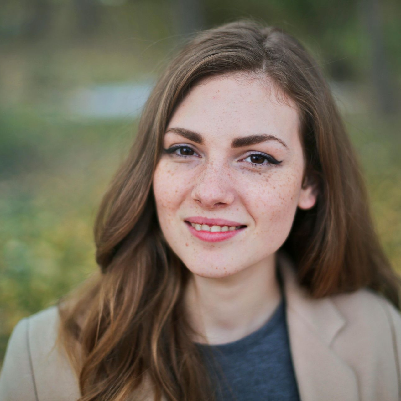 Lachende vrouw met lang bruin haar in een beige jas tegen een wazige buitenachtergrond.
