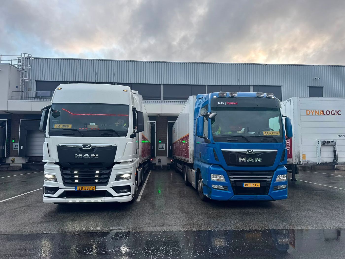 Twee geparkeerde MAN-vrachtwagens, een witte en een blauwe, voor een magazijn op een regenachtige dag.