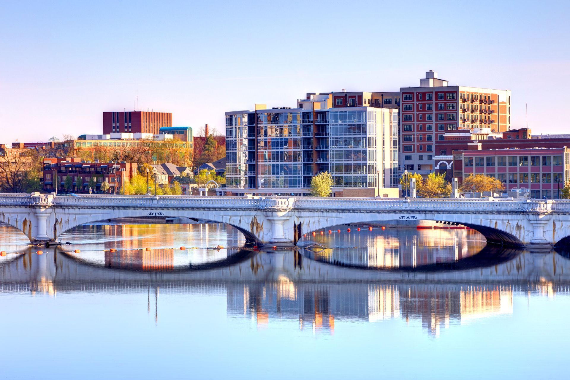 Handshake between a person in a blue jumpsuit holding a toolbox and a person wearing jeans and a light-colored top, by a doorway. City skyline reflected in water, arched bridge in foreground. Buildings of various heights under blue sky.