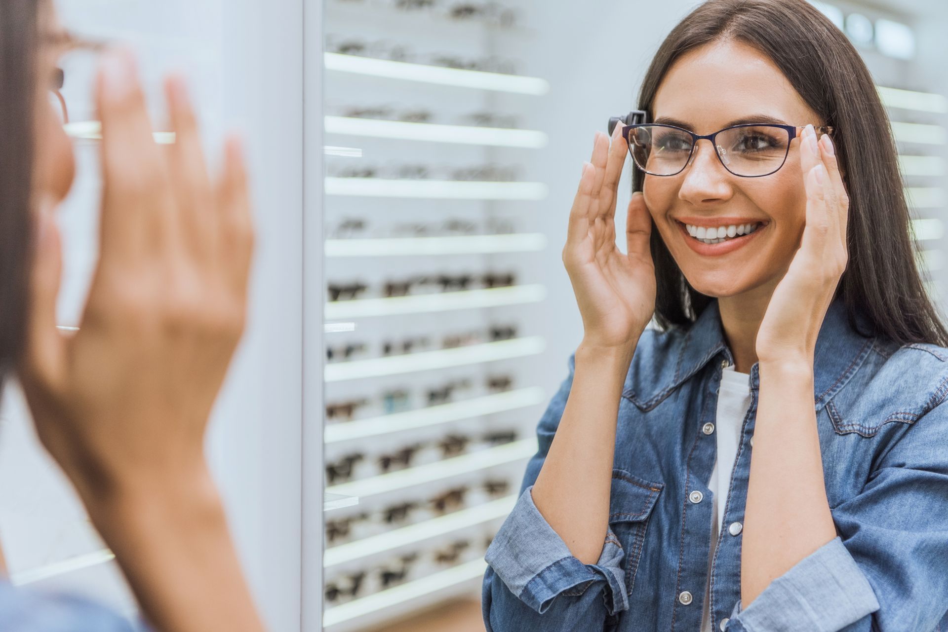 A smiling woman choosing eyeglasses and looking at a mirror in an optical