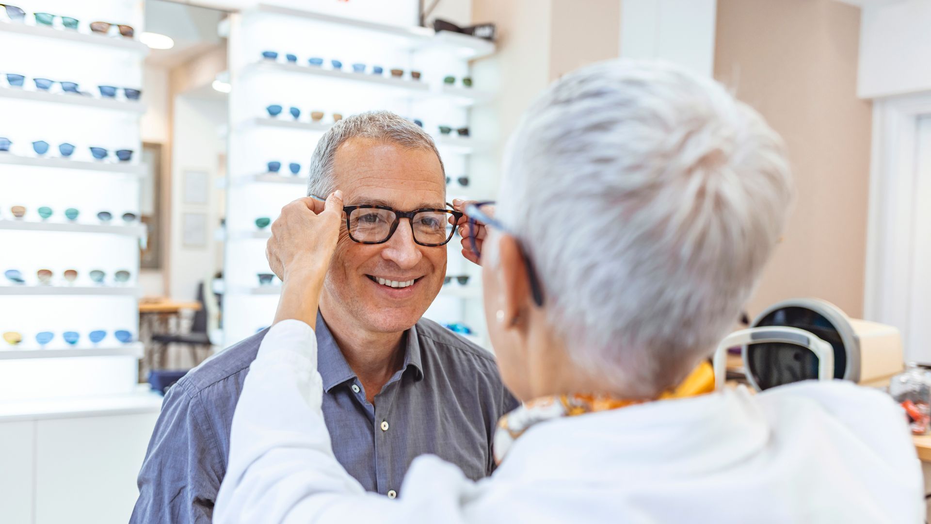 A woman is helping a man try on glasses in an optical shop.