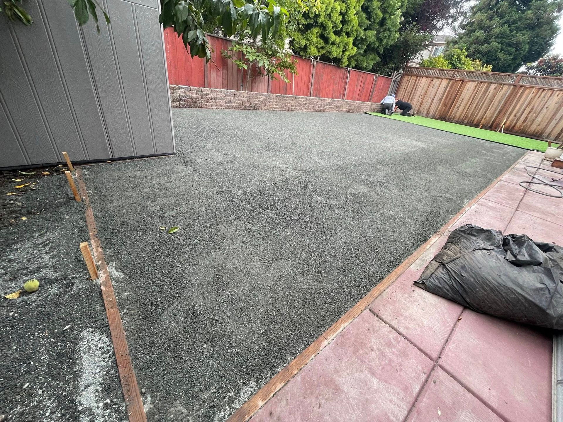 Backyard with freshly laid gravel, red fence, and a black bag on a pink patio edge