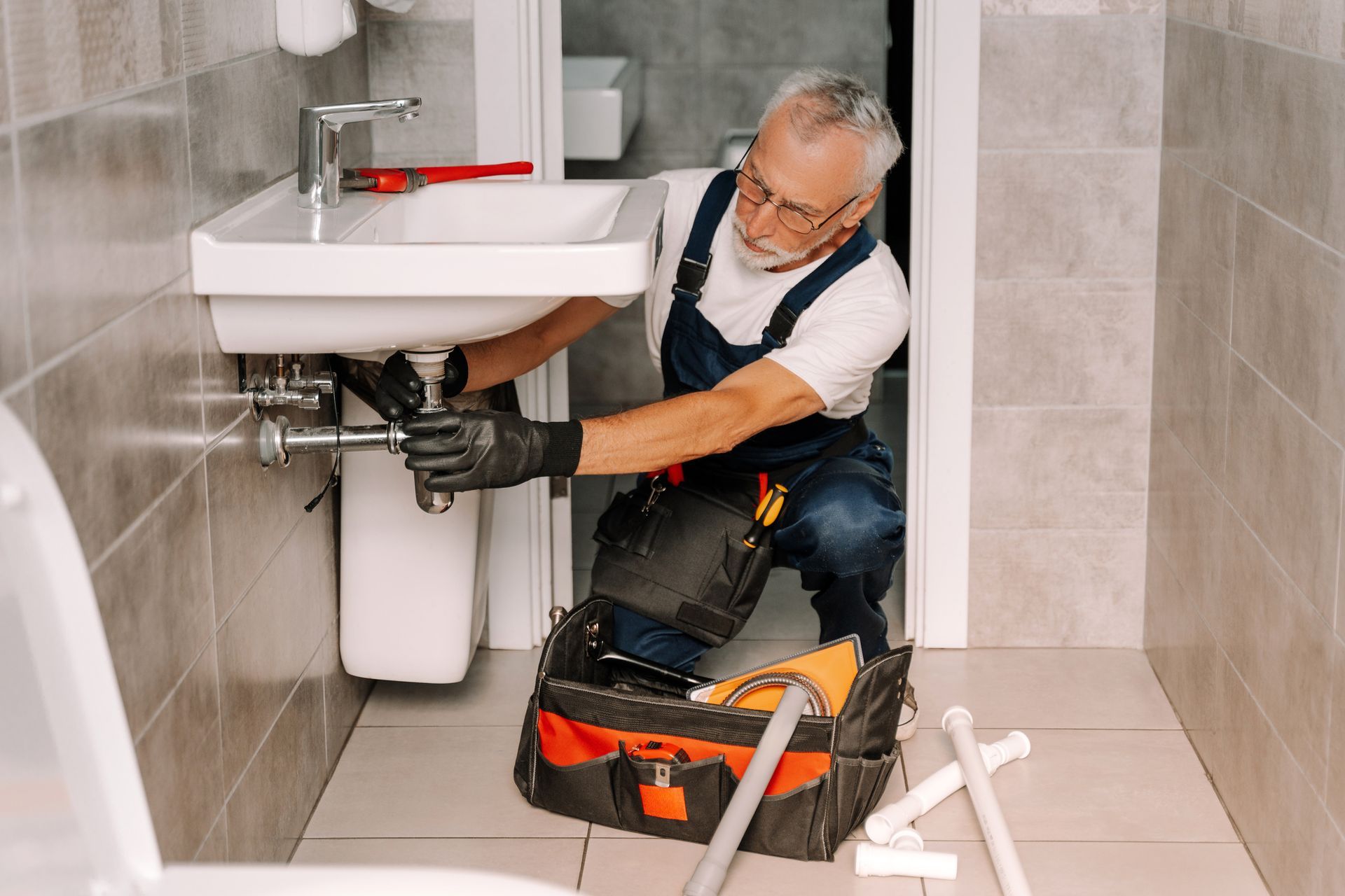 A plumber under the bathroom sink installing piping with a wrench, with a toolbox nearby.