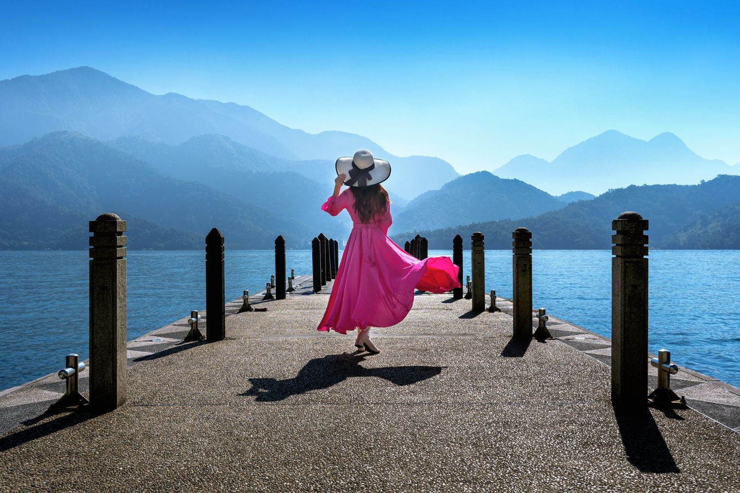 A woman in a pink dress and hat is standing on a pier overlooking a lake.
