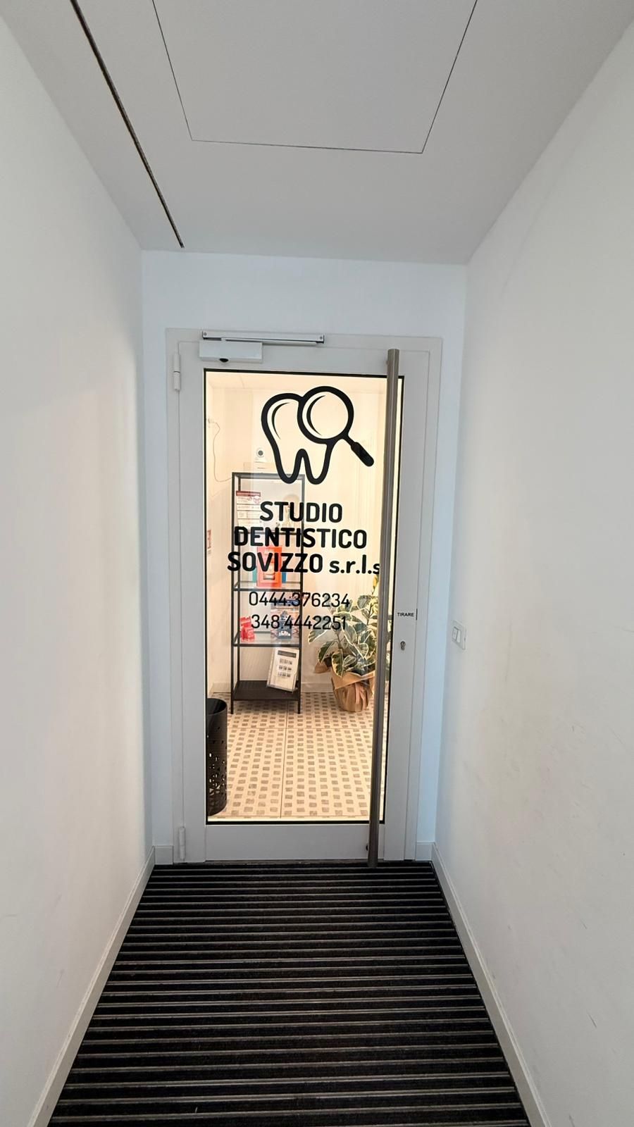 Stretto corridoio bianco che conduce a una porta a vetri con l'insegna dello studio e un logo circolare.