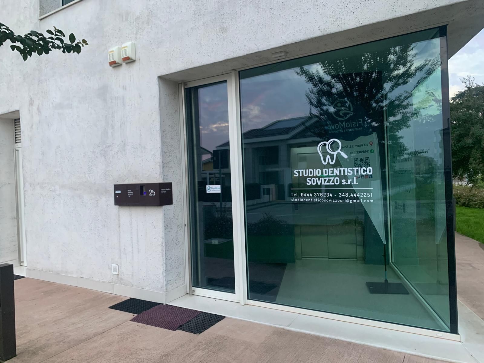 Vetrina d'ingresso con logo e testo bianco sulla porta, accanto a una parete esterna bianca.