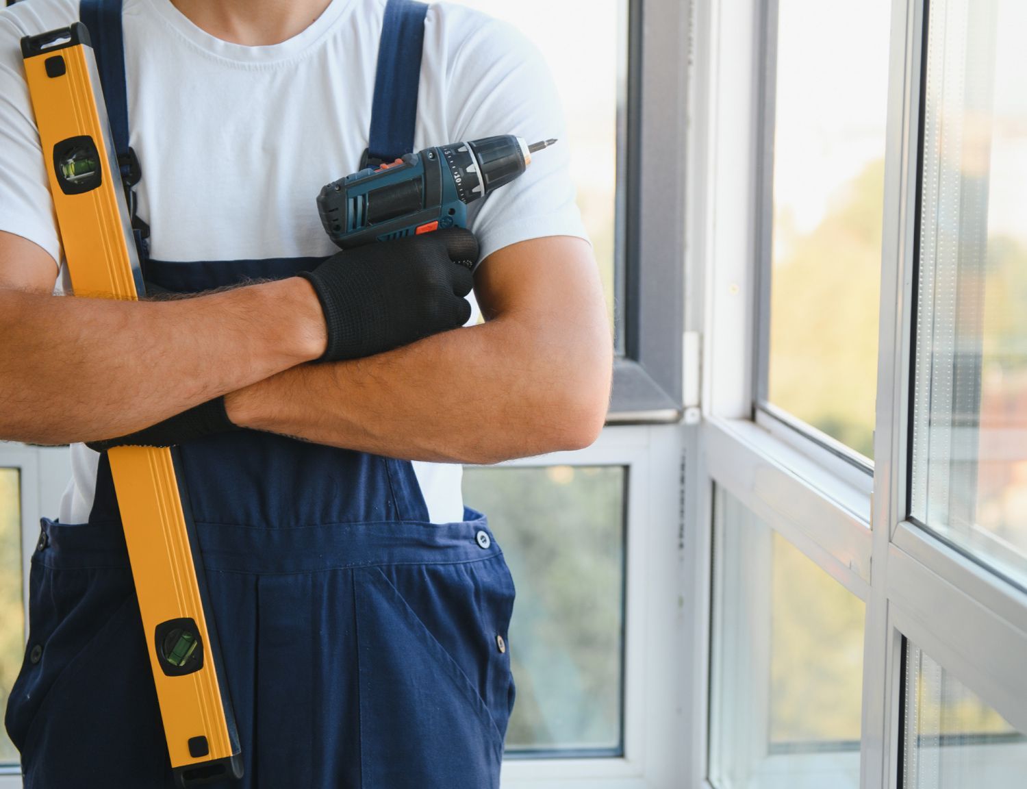 A person in overalls stands by a window, holding a yellow spirit level and a power drill A person in overalls stands by a window, holding a yellow spirit level and a power drill