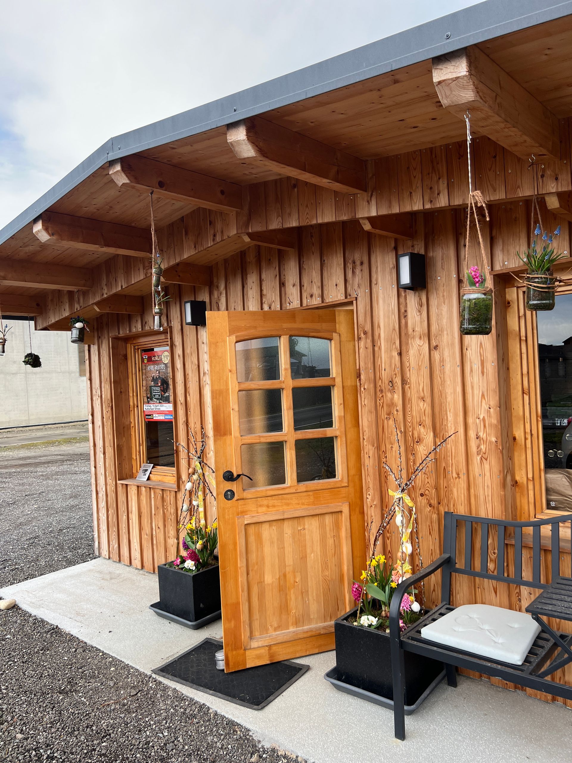 Eingang zur Holzhütte mit offener Tür, hängenden Blumen und einer Bank auf einer kleinen Veranda