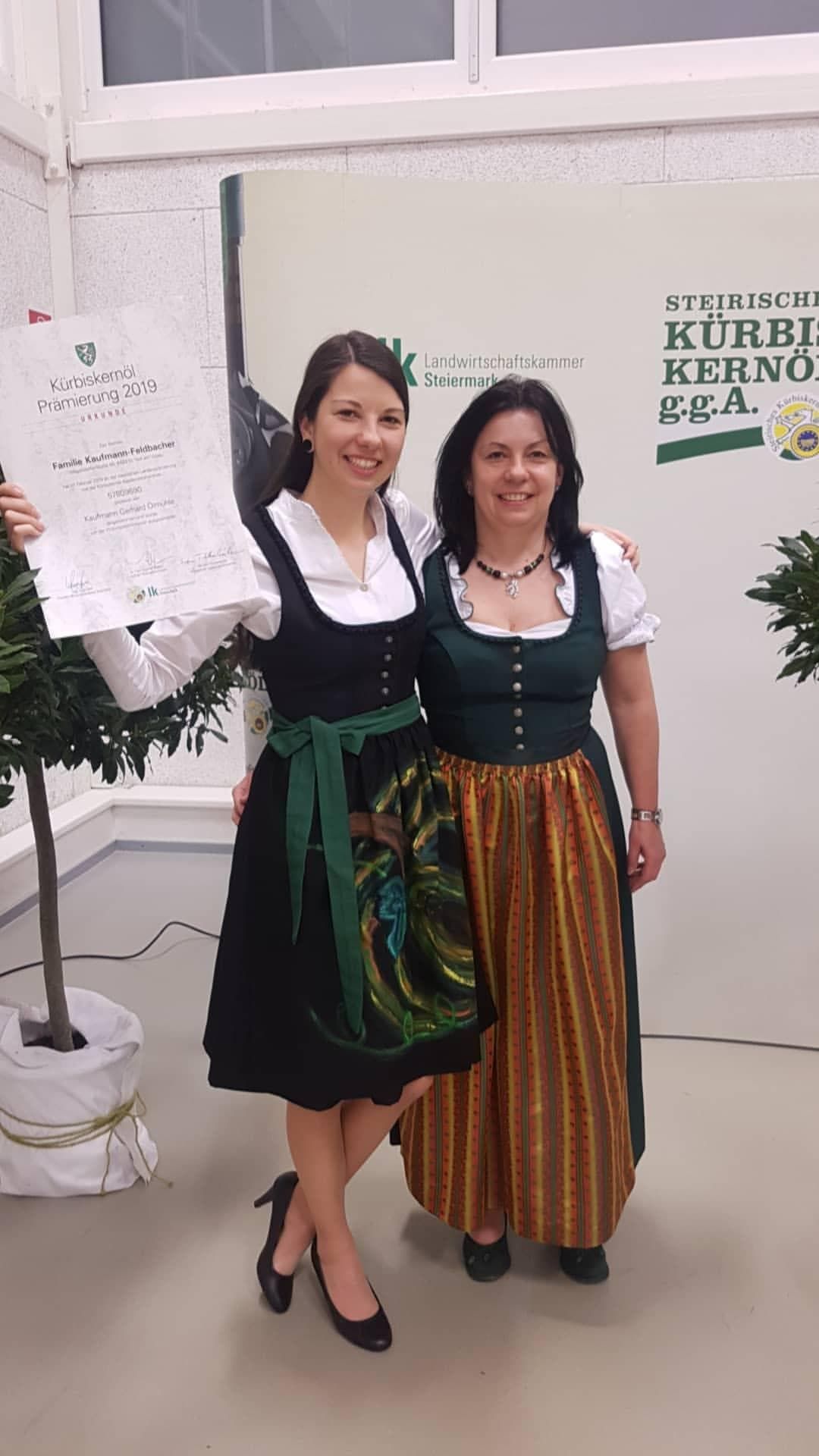 Zwei Frauen in traditioneller Tracht stehen lächelnd vor einer Veranstaltungskulisse.