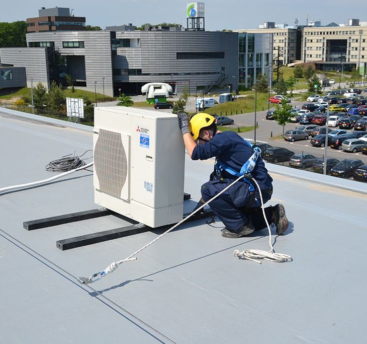 A man wearing a yellow helmet is working on an air conditioner on the roof of a building